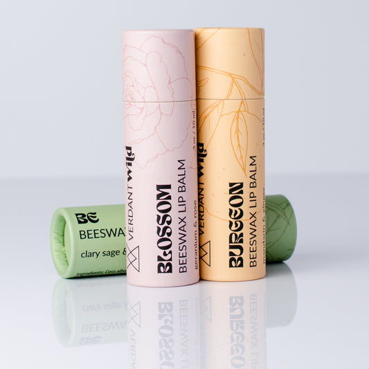 Three cylindrical tubes of Verdant Wild beeswax lip balm arranged on a reflective white surface against a light gray studio background. The center tube is pale pink labeled BLOSSOM, the right tube is pale yellow-orange labeled BURGEON, and a green tube labeled CLARY SAGE lies on its side in the foreground.