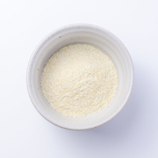 A top-down view of a small off-white ceramic bowl filled with Verdant Wild Brighten Facial Grains (chamomile & turmeric): a fine, pale yellow powder, set against a clean white background.