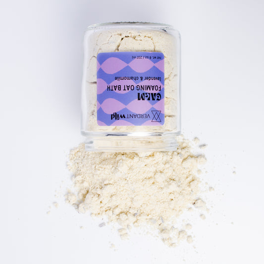A clear glass jar filled with a powdered milk bath, featuring a purple and lavender patterned label. Some of the beige powder is spilled in a small pile in front of the jar on a clean white background.