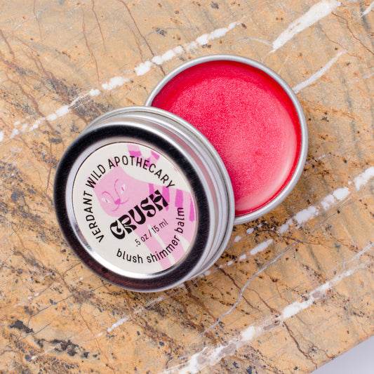 An open round metal tin of blush shimmer balm sits on a beige stone surface. The balm inside is a bright pink shimmer.