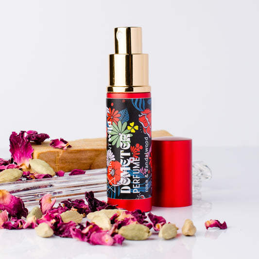 A perfume atomizer bottle with a gold pump and a colorful floral label, shown with its red cap beside it on a white surface. Dried rose petals, cardamom and sandalwood are styled around the product.