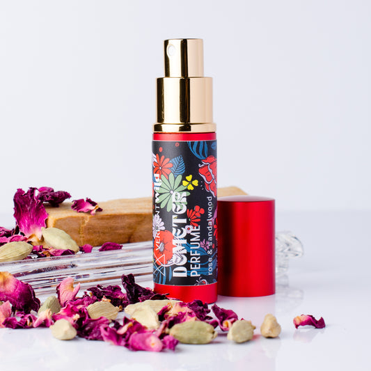 A perfume bottle labeled 'DEMETER PERFUME' with a colorful floral pattern, gold pump, and red cap, styled with dried rose petals, cardamom pods, and a wooden block on a white background.