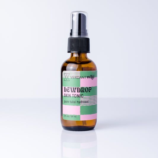 A 2 fl oz amber glass spray bottle of Verdant Wild Tulsi Skin Tonic with a black spray cap and a pastel pink, green, and white label, set against a clean white background with a soft reflection.