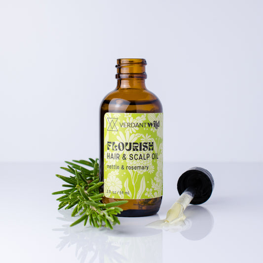 An amber glass bottle of Verdant Wild Flourish Hair & Scalp Oil with a green and white label, shown on a white reflective surface with a green rosemary sprig and a black dropper lying beside it.