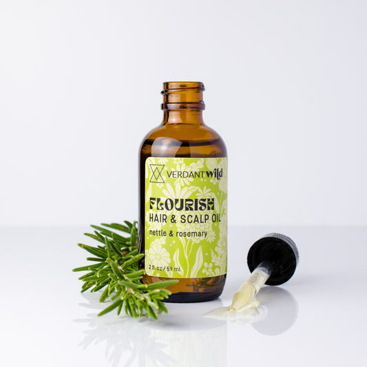 An amber glass bottle of Verdant Wild Flourish Hair & Scalp Oil with a light green and white patterned label, shown with a green rosemary sprig and a black dropper lying on its side on a reflective white surface.