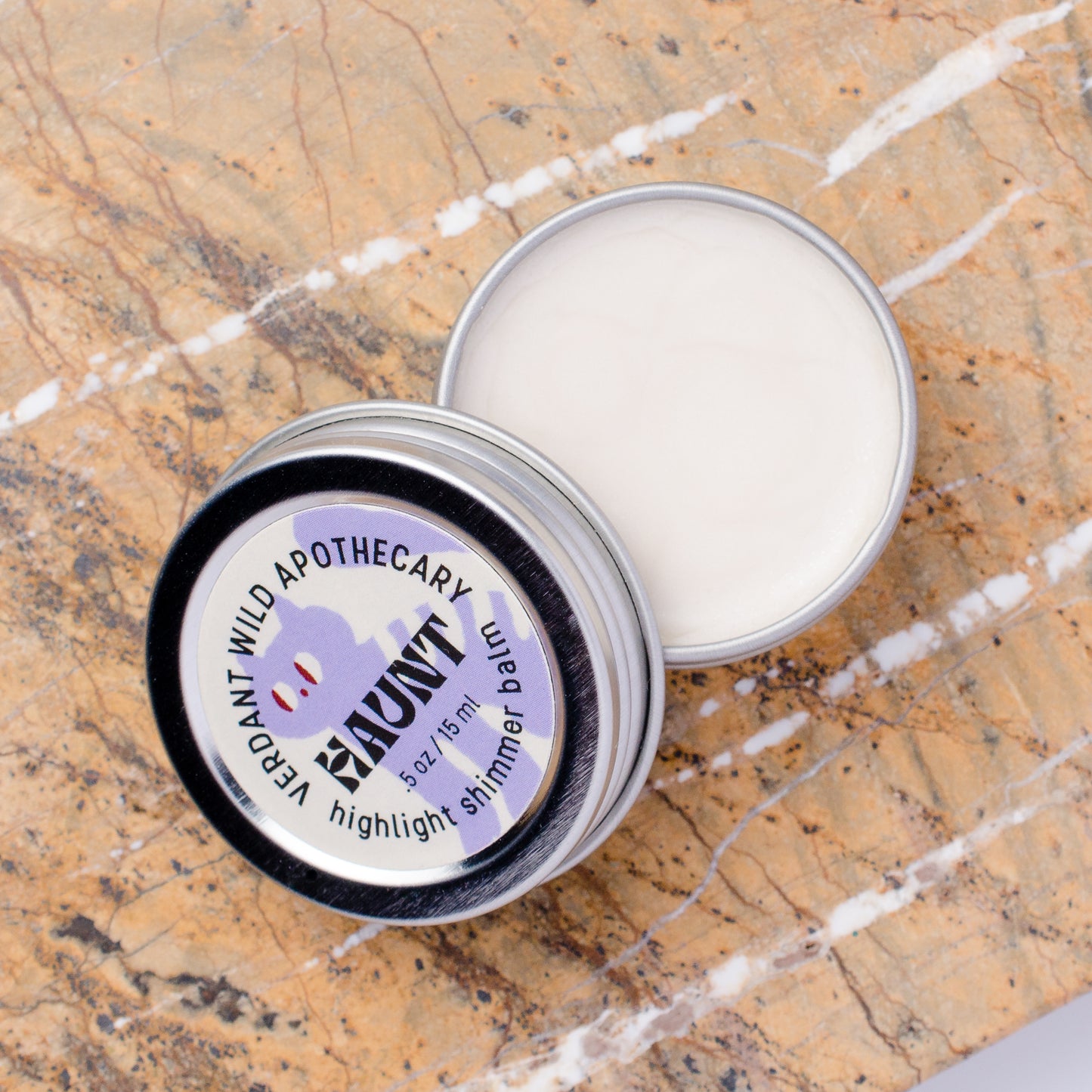 An open silver metal tin of highlighter shimmer balm lies on a brown and white marble surface, with the lid placed beside it.