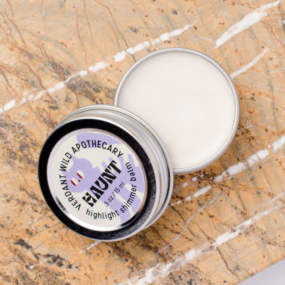 An open round metal tin of highlighter balm sits on a beige marble surface, with the lid placed beside it. The label reads 'HAUNT' and the cream inside is visible.