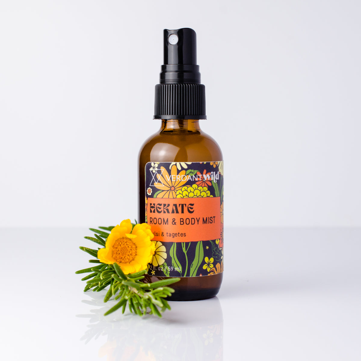 An amber glass spray bottle labeled 'HEKATE ROOM & BODY MIST' with a black pump, standing on a white surface against a light gray background, with a yellow flower and green sprigs placed in front.