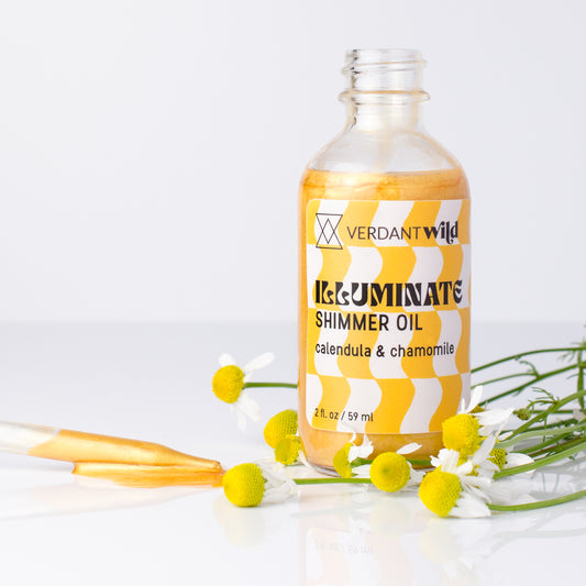 A clear glass bottle of Verdant Wild Illuminate Shimmer Oil with a yellow and white patterned label, styled with chamomile flowers and a glass pipette on a white reflective surface.