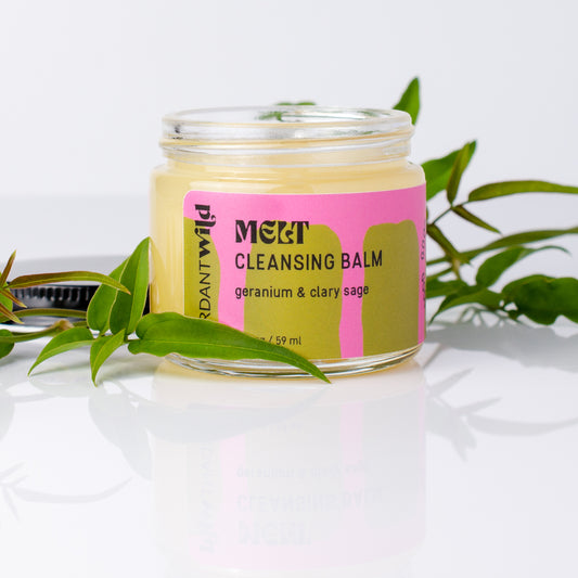 A glass jar of MELT Cleansing Balm with a pink and olive-green label, styled with green leaves on a reflective white surface.