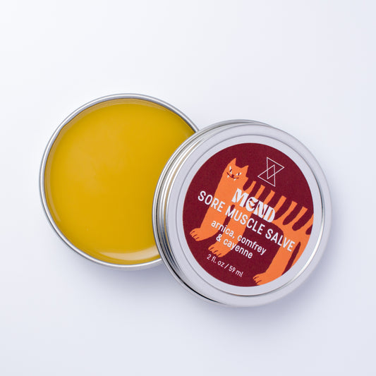 An open round metal tin of Mend Sore Muscle Salve showing the yellow balm inside, with the lid leaning against it. The lid features a dark red label with an orange cat illustration and white text.