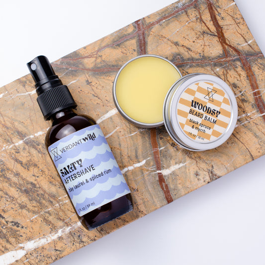 A grooming set featuring a dark amber spray bottle of Verdant Wild Salty Aftershave and a round tin of Woodsy Beard Balm, arranged on a brown and white marble slab against a white background.