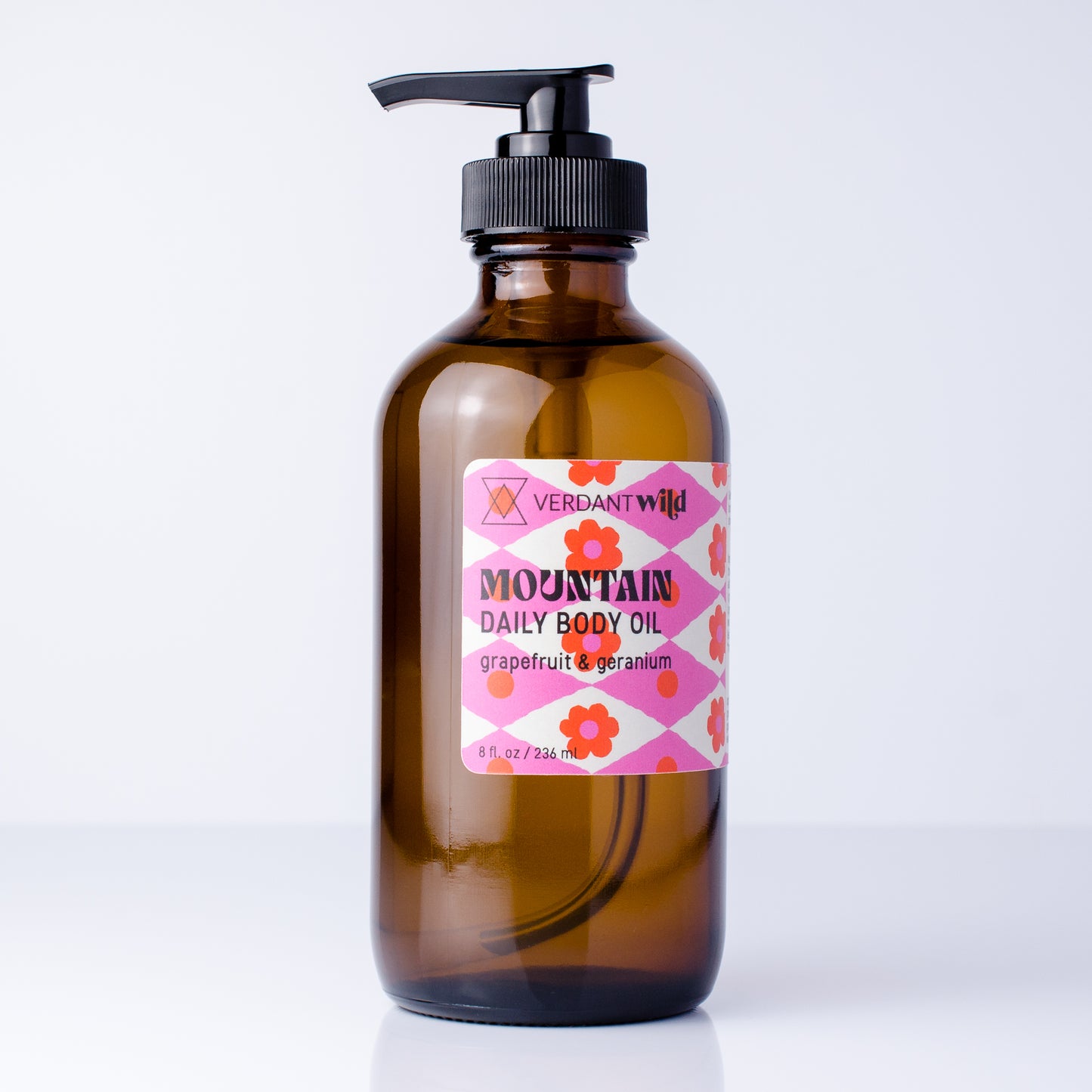 An amber glass pump bottle of Verdant Wild Mountain Daily Body Oil with a pink and orange floral label, set against a clean white background.