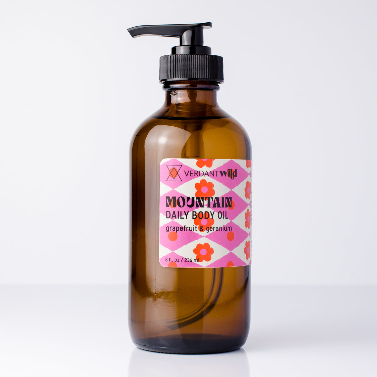 An amber glass pump bottle of Verdant Wild Mountain Daily Body Oil with a pink and orange floral label, set against a clean white background.
