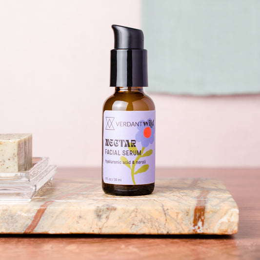 A small amber glass pump bottle labeled 'Verdant Wild Nectar Facial Serum' stands on a marble slab. The bottle has a black pump and a lavender label with floral graphics.
