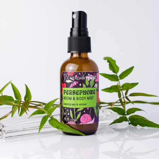 A bottle of Persephone Room & Body Mist with sweetgrass and vetiver scent, styled with green leaves on a reflective white surface against a white background.