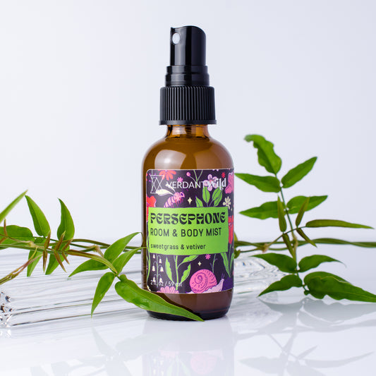 An amber glass spray bottle labeled 'Verdant Wild Persephone Room & Body Mist' with a black pump, styled with green leaves on a reflective white surface against a light gray background.