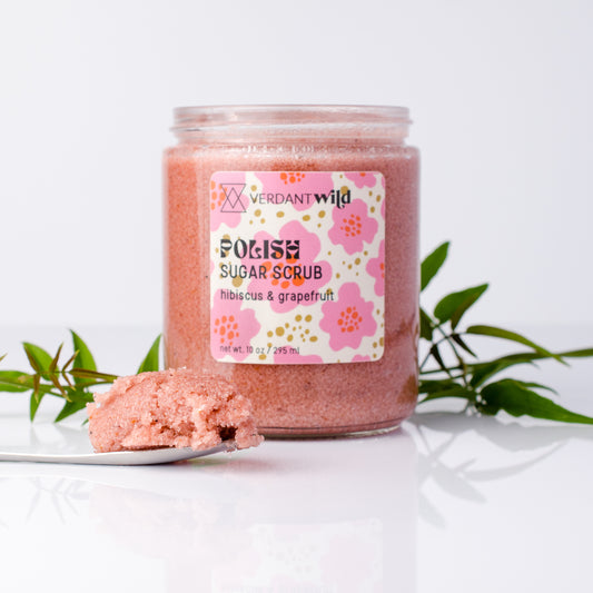 A jar of Verdant Wild Sugar Scrub with a pink hibiscus and grapefruit scent, displayed on a white surface with a scoop of the scrub and green leaves in the background.