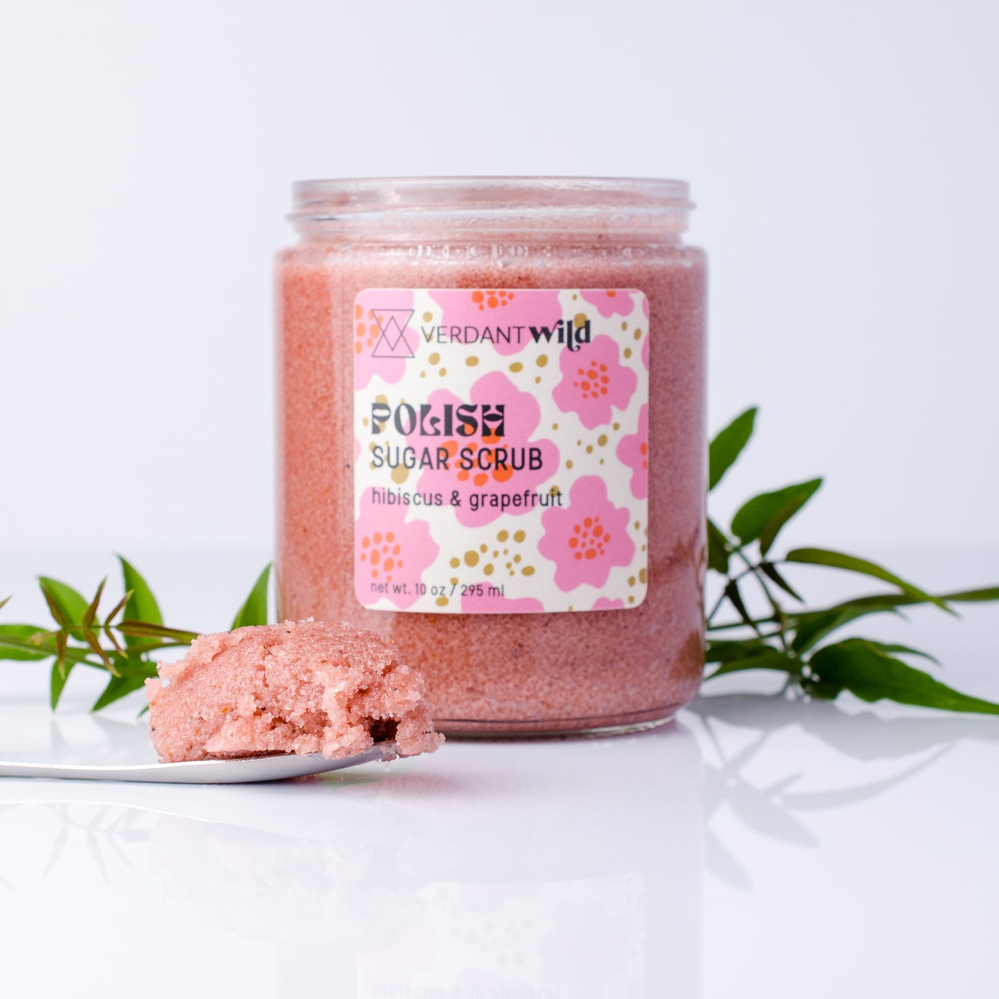 A jar of Verdant Wild Soak Sugar Scrub with hibiscus and grapefruit, displayed on a reflective white surface with a scoop of scrub and green leaves in the background.
