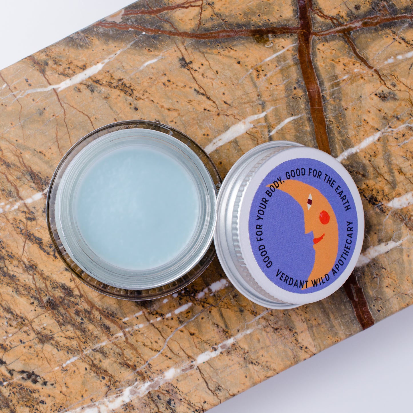 An open round glass jar containing a pale blue cream, placed on a brown marbled surface next to the silver lid. The lid has a colorful label with a crescent moon illustration and the text 'GOOD FOR YOUR BODY, GOOD FOR THE EARTH VERDANT WILD'.