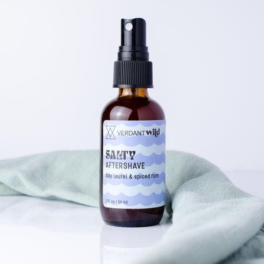 A 2 fl oz amber glass spray bottle of Verdant Wild Salty Aftershave with a black pump, set on a white surface with a light sage green towel in the background.