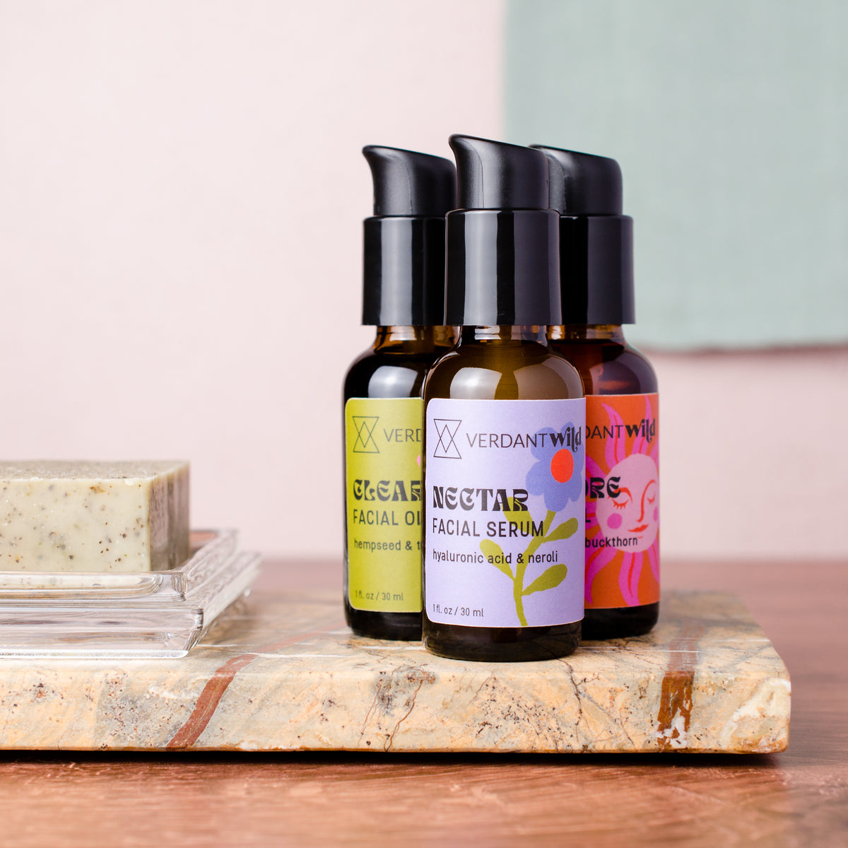 Three small cosmetic bottles with black pumps are arranged on a stone slab beside a bar of soap. The center bottle is labeled 'VERDANT WILD NECTAR FACIAL SERUM,' with two additional bottles behind it partially visible.