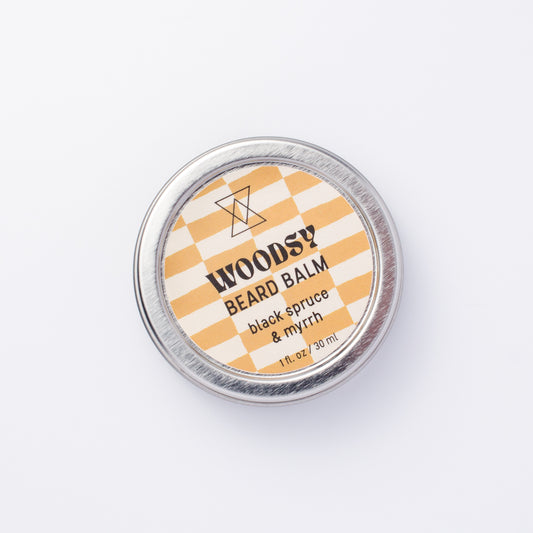 A round silver tin of WOODSY Beard Balm in the 'black spruce & myrrh' scent, featuring a yellow and white checkered label, photographed from directly above on a clean white background.