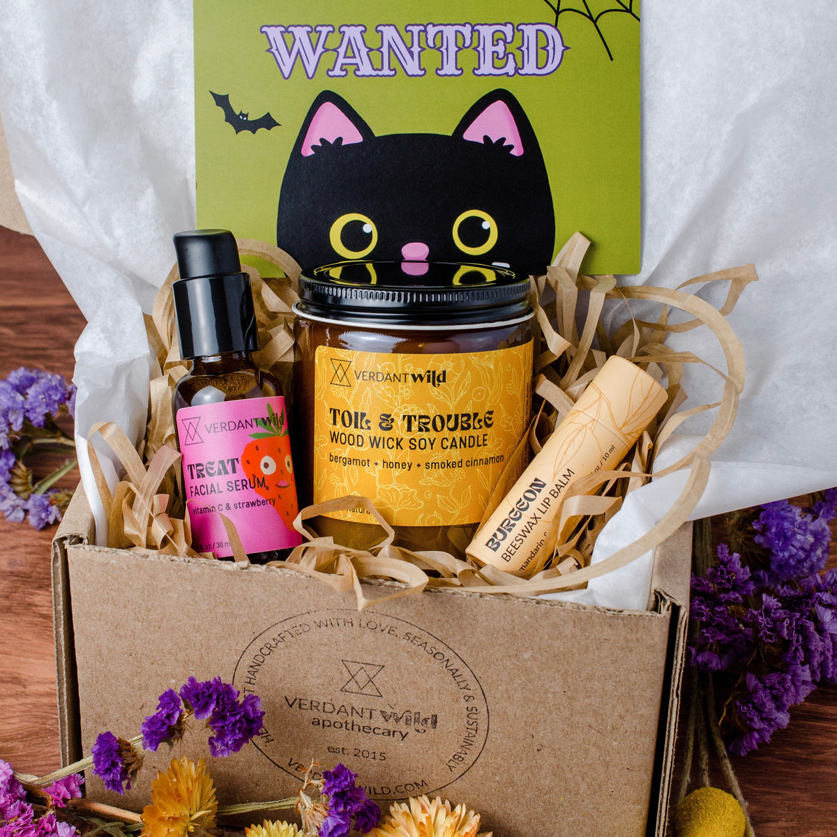 A spooky-themed apothecary box from Verdant Wild, containing a 'Treat vitamin C Facial Serum' bottle, a 'Toil & Trouble' wood wick soy candle, and a 'Burgeon' lip balm, arranged in a kraft box with shredded paper and a green 'WANTED' cat card.