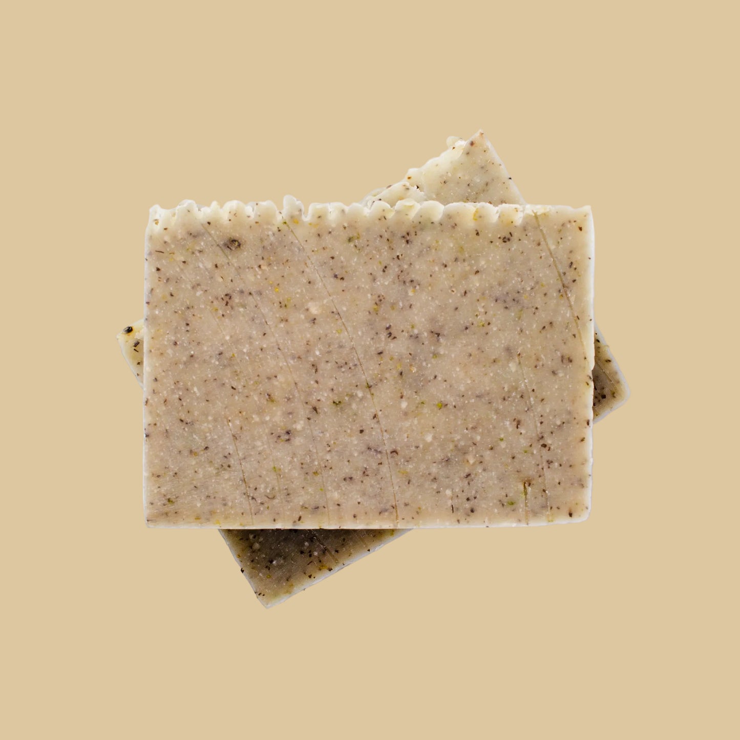 Two rectangular bars of handmade soap stacked on a neutral background, with the top bar showing a speckled beige and green texture.