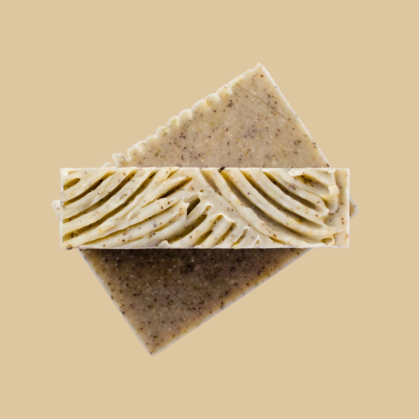 Two rectangular bars of handmade soap stacked diagonally on a clean beige background. The top bar shows a decorative ridged cut surface, while the sides are olive green and speckled.