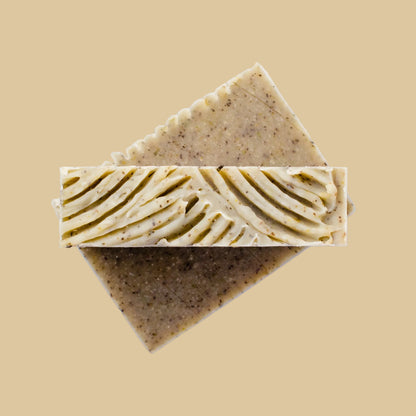 Two rectangular bars of handmade soap stacked diagonally on a clean beige background. The top bar shows a decorative ridged cut surface, while the sides are olive green and speckled.