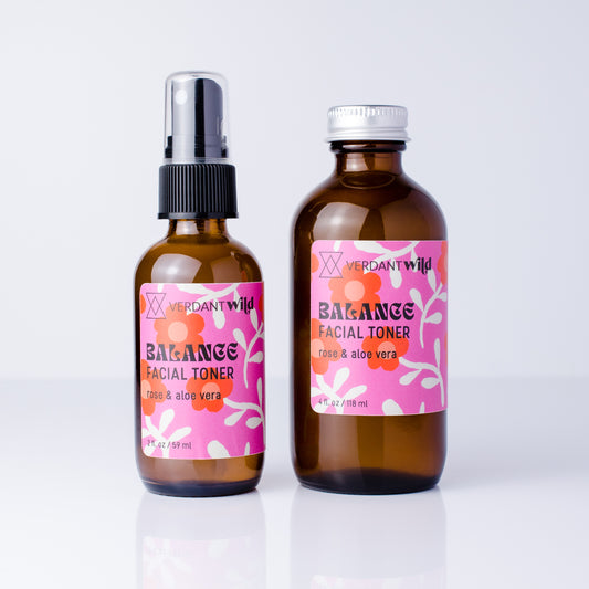 Two amber glass bottles of Verdant Wild Balance Facial Toner with pink floral labels, one with a black spray top and one with a silver cap, on a white reflective surface.