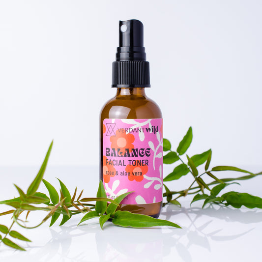 A bottle of Verdant Wild Balance Facial Toner with a pink label and black spray cap, styled with green leaves on a clean white background.