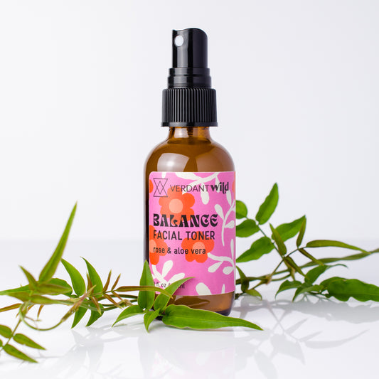 A bottle of Verdant Wild Balance Facial Toner with a pink floral label and black spray cap, styled with green leaves on a clean white background.
