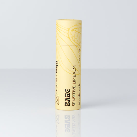 A single tube of Bare Sensitive Lip Balm in a pale yellow color with black text and a botanical line drawing, standing upright on a reflective light gray surface.