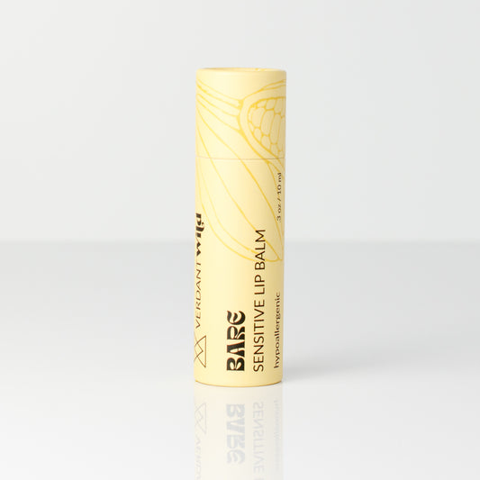 A single tube of Bare Sensitive Lip Balm in a pale yellow color with gold line art and black text, standing upright on a reflective white surface against a white background.