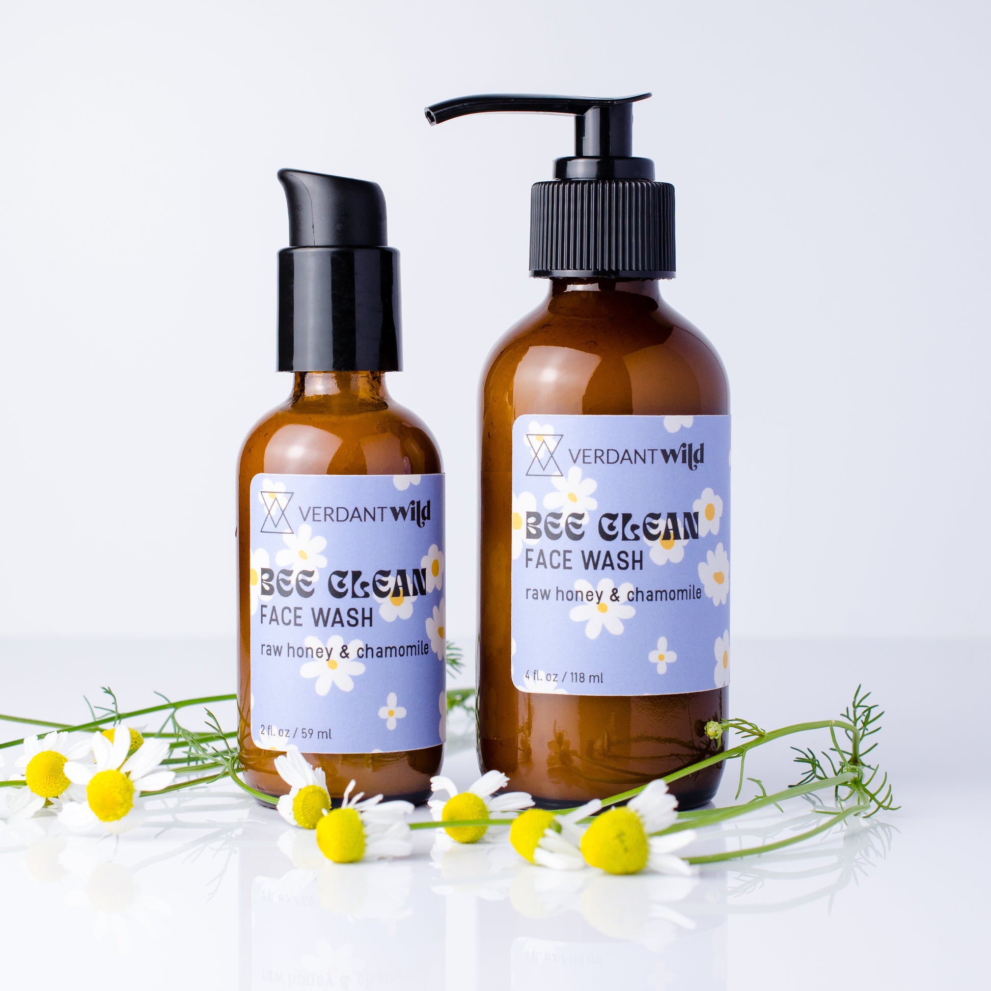 Two amber glass face wash bottles with black pumps and lavender labels reading Verdant Wild BEE CLEAN FACE WASH, styled with chamomile flowers on a white background.