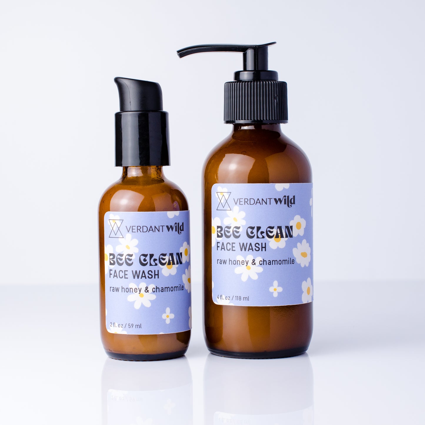 Two amber glass face wash bottles with black pumps on a white reflective surface. The larger bottle has a black pump and the smaller one has a black cap, both with lavender labels featuring white daisy flowers and the brand name Verdant Wild.