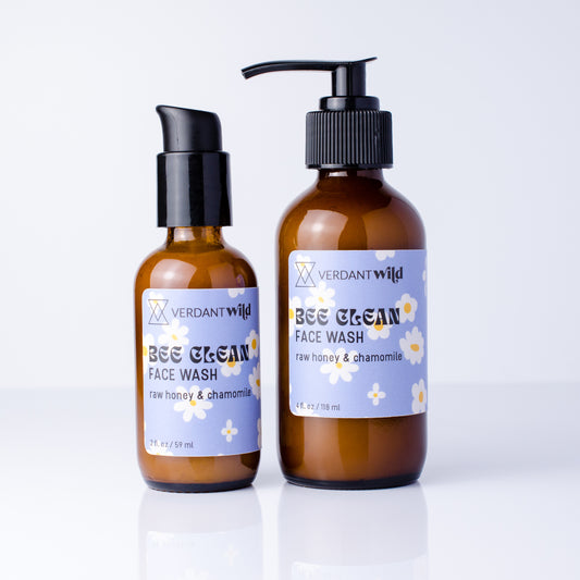 Two amber glass face wash bottles with black pumps on a white reflective surface. The larger bottle has a black pump and the smaller one has a black cap, both with lavender labels featuring white daisy flowers and the brand name Verdant Wild.