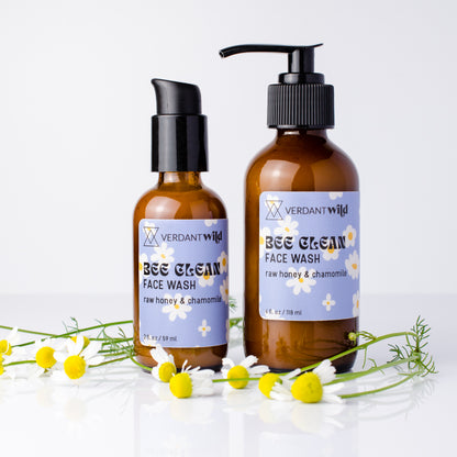 Two amber glass bottles of Verdant Wild Bee Clean Face Wash, decorated with fresh chamomile flowers on a white reflective surface.