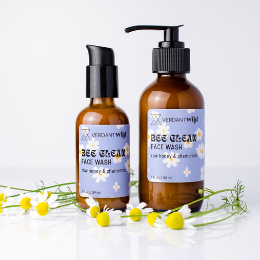 Two amber glass bottles of Verdant Wild Bee Clean Face Wash, decorated with fresh chamomile flowers on a white reflective surface.
