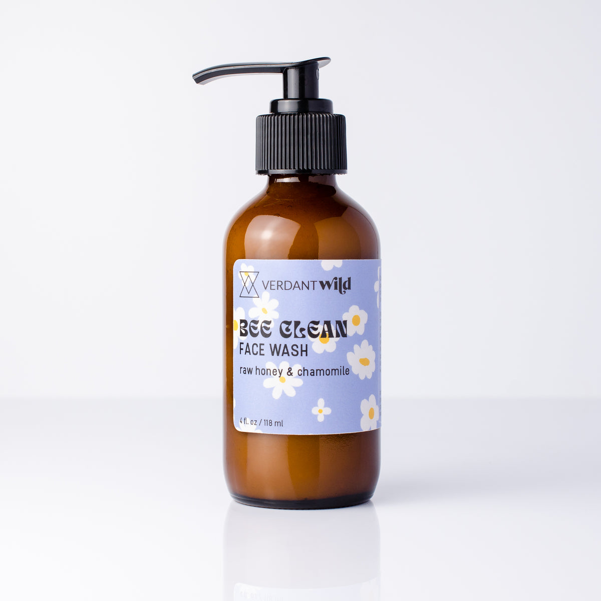 A 4 fl oz amber glass bottle of Verdant Wild Bee Clean Face Wash with a black pump dispenser and a light blue label decorated with white chamomile flowers, set against a clean white background.