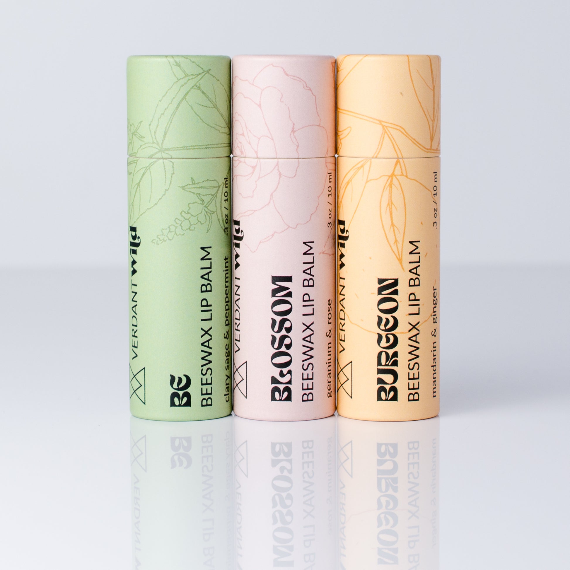 Three vertical tubes of Verdant Wild beeswax lip balm in different colors: sage green, pale pink, and peach, arranged side by side on a reflective light gray surface.