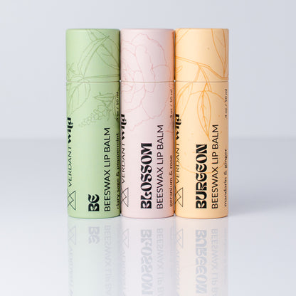 Three vertical tubes of Verdant Wild beeswax lip balm in different colors: sage green, pale pink, and peach, arranged side by side on a reflective light gray surface.