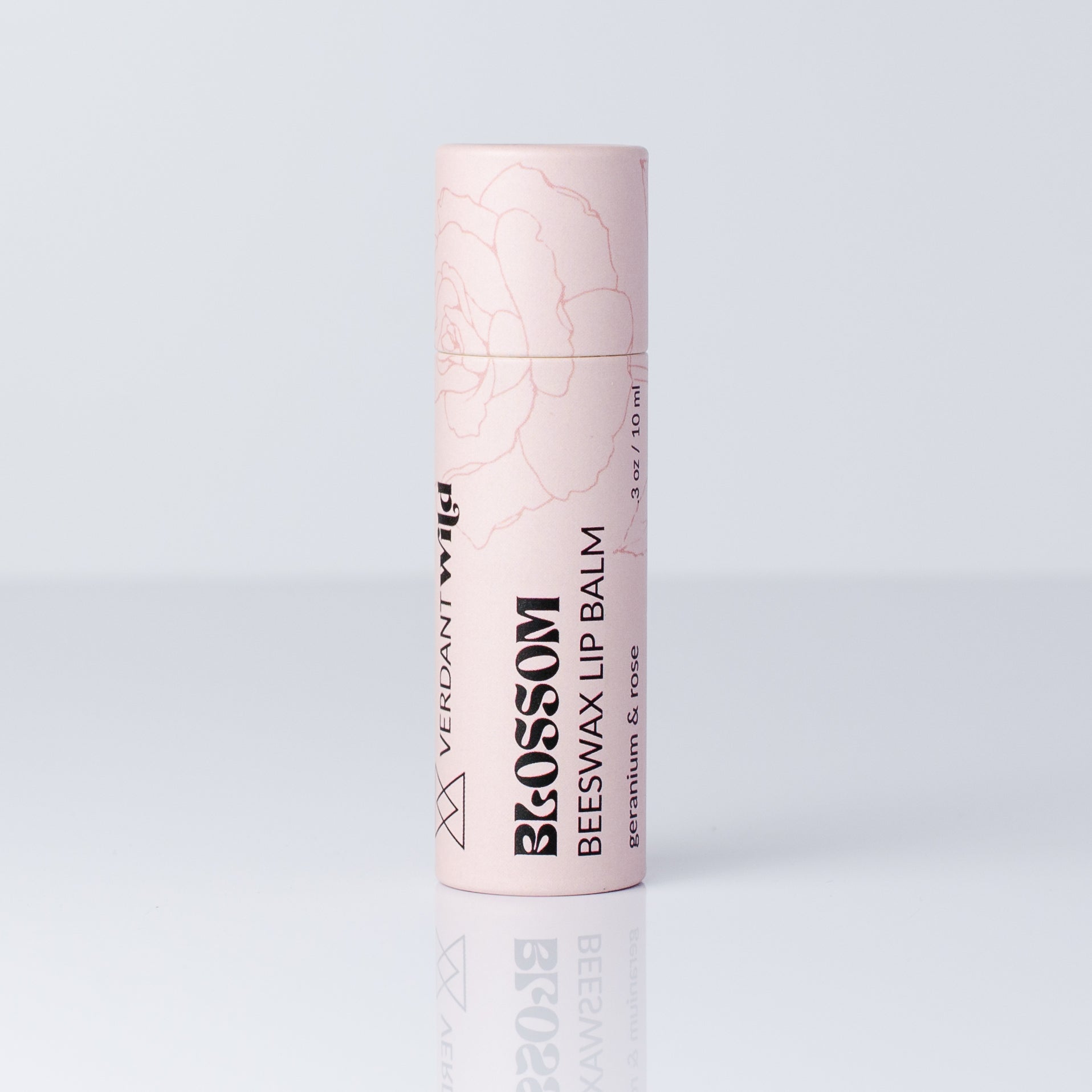 A single tube of Verdant Wild Blossom Beeswax Lip Balm in a pale pink color with black text and a faint rose line drawing, set against a light gray background with a soft reflection below.