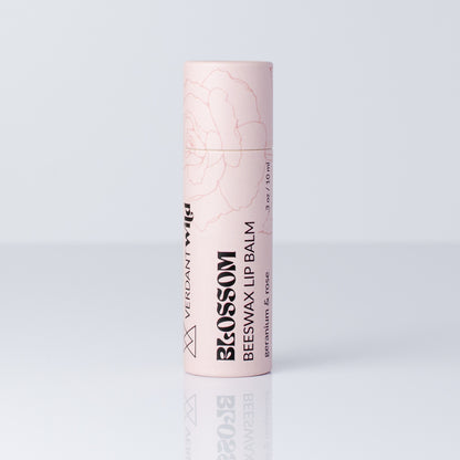 A single tube of Verdant Wild Blossom Beeswax Lip Balm in a pale pink color with black text and a faint rose line drawing, set against a light gray background with a soft reflection below.