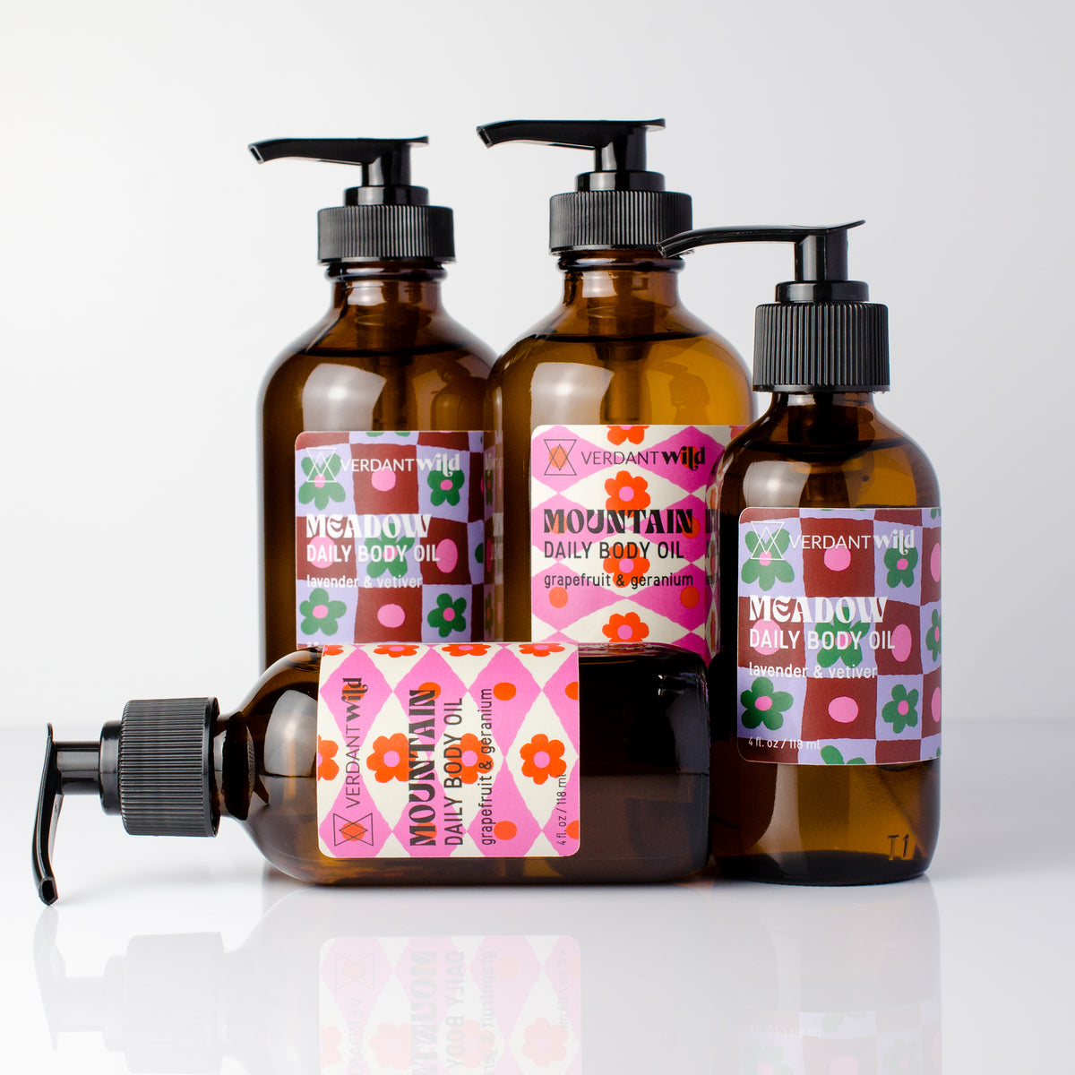 Four amber glass pump bottles of VERDANT WILD Daily Body Oil with black pumps and colorful patterned labels, arranged on a white reflective surface.