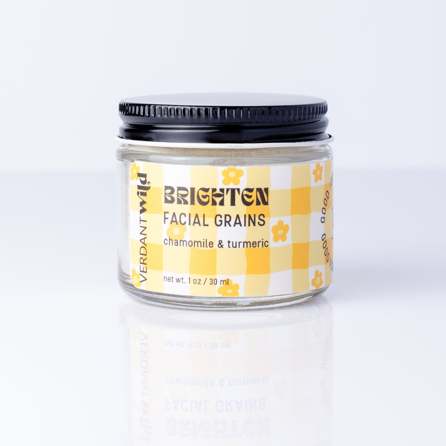 A small glass jar of Verdant Wild Brighten Facial Grains (chamomile & turmeric) with a black lid and a yellow-and-white checkered label, set against a clean white background with a soft reflection.