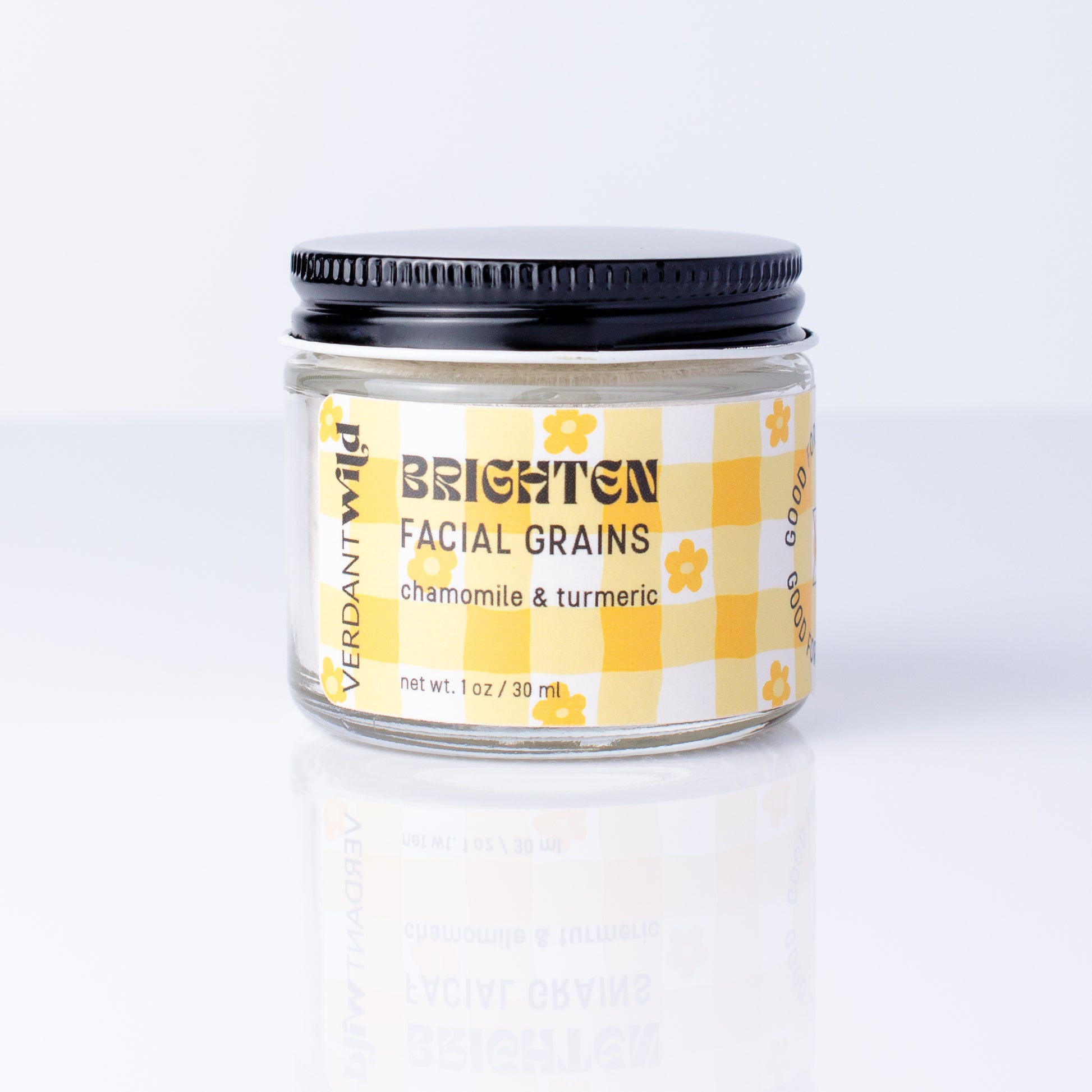 A small glass jar of Verdant Wild Brighten Facial Grains (chamomile & turmeric) with a black lid and a yellow-and-white checkered label, set against a clean white background with a soft reflection.