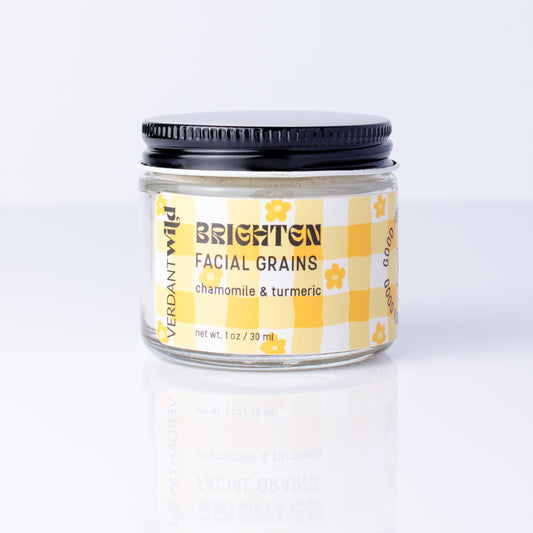 A small glass jar of Verdant Wild Brighten Facial Grains (chamomile & turmeric) with a black lid and a yellow-and-white checkered label, set against a clean white background with a soft reflection.
